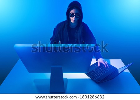Hacker computer. Computer and laptop in front of a hacker. Guy in hood is working with a  laptop. Hacker in neon light. Concept - man makes attack on computer networks. Breaking. Hacking.