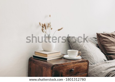 Modern white ceramic vase with dry Lagurus ovatus grass and cup of coffee on retro wooden bedside table. Beige linen and velvet pillows in bedroom. Scandinavian interior. Homestaging. Royalty-Free Stock Photo #1801312780