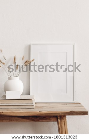 Portrait white picture frame mockup on vintage bench, table. Modern ceramic vase with dry Lagurus ovatus grass, books and busines card. White wall background. Scandinavian interior. Vertical.