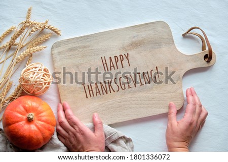 Hands hols cutting board with text Happy Thanksgiving. Fall decorations, golden wheat ears and orange pumpkin. Top view, flat lay on table with white textile tablecloth.