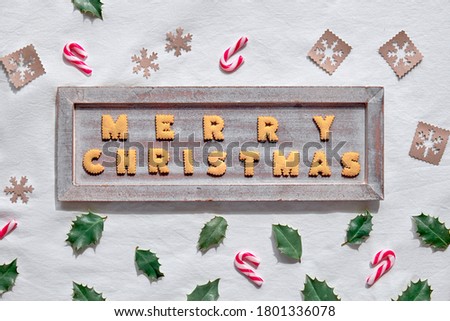 ext Merry Christmas made with cookies on rustic wooden board. Geometric flat lay on white textile background. Eco-friendly decorations from craft paper , candy canes and natural holly leaves.