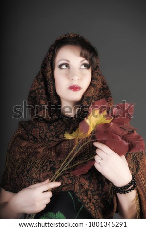 beautiful Russian brunette woman with a scarf on her head and autumn leaves in her hands, photo in the studio on a gray background