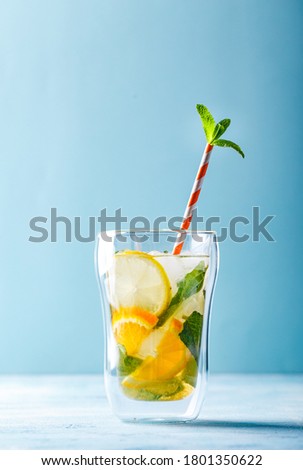 Summer refreshing cocktails, lemonade or mojito with lemon, orange and mint with ice in a glass cup and with a striped paper straw. On a light blue background. Copy space.