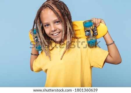 Happy little boy with african dreads holding skateboard on shoulders and smile over blue background.