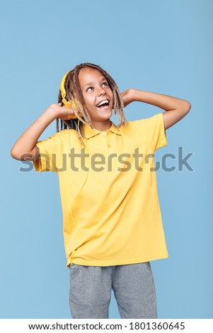 Joyful little boy with african dreads listening music in headphones has pleasant smile over blue background.