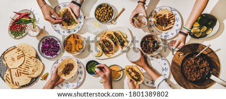 Friends having Mexican Taco dinner. Flat-lay of beef tacos, tomato salsa, tortillas, beer, snacks and peoples hands over white table, top view. Mexican cuisine, gathering, feast, comfort food concept Royalty-Free Stock Photo #1801389802