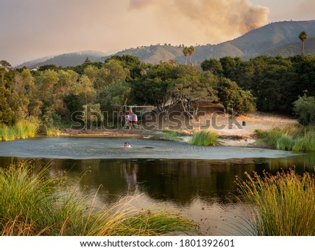 A helicopter pilot collects a bucket of water from a pond to douse the "River Fire" in the hills Monterey County of the central coast of California as plumes of smoke drift upward.     Royalty-Free Stock Photo #1801392601