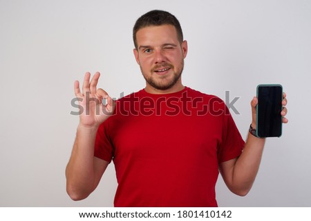 Young handsome Cucasian man wearing red shirt standing against white background holding in hands cell showing ok-sign  Royalty-Free Stock Photo #1801410142