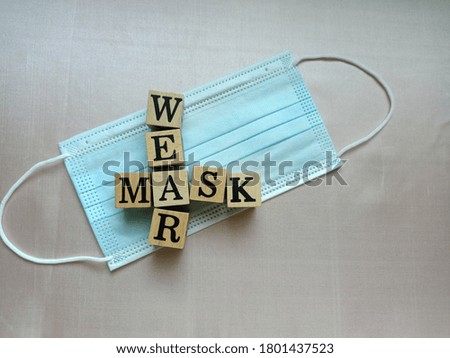 Top view photo of the wooden square block with the words WEAR MASK on a disposable mask, isolated on beige silk cloth for decorative design and advertising.