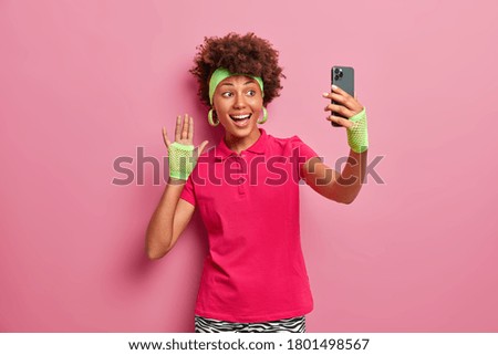 Positive sportswoman in active wear waves hand at camera of smartphone, takes selfie, sends picture to followers, has happy mood, makes greeting gesture, smiles at mobile display, poses indoor