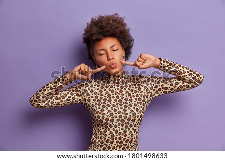 Portrait of dark skinned relaxed woman with curly hair, closes eyes, points index fingers at cheeks, keeps lips folded, makes grimace, dressed in leopard turtleneck, isolated over vivid purple wall
