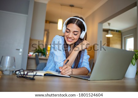 A young happy college female student sitting at the table at home, using tablet and headphones when studying. Focused woman wearing headphones watching webinar write notes