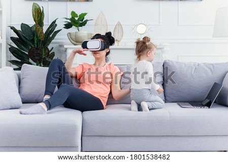Pretty woman uses virtual reality goggles at home on sofa while her daughter watches cartoons.. Plays games, technologies of the future.