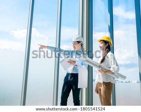 Asian architect Male and female inspecting construction work inside high-rise building near huge window glass on blue sky background. Couple engineer in hard hats holding blueprint in new building.