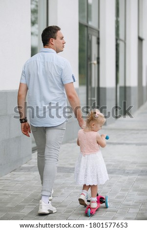Father walks with his daughter holding hands along the city street. A blonde girl with ponytails is rolling on a scooter. Back view.