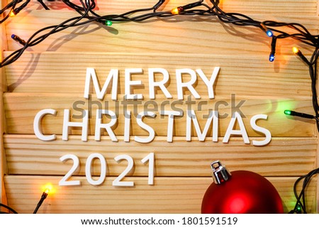 White letters text Merry Christmas 2021 on a wooden background with a garland of colored lights.Christmas tree decoration red ball.Lettering on the background.Happy New Year!christmas card. Flat lay