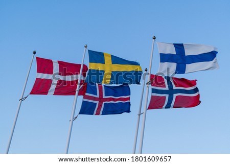 Scandinavian flags waving on top of metal poles against blue sky.