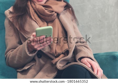 Crop female in warm brown coat and scarf interacting with smartphone while sitting in soft armchair in corridor of contemporary high rise building