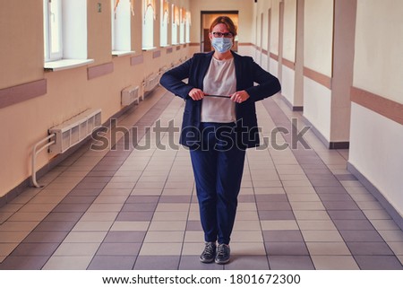 An angry teacher in a medical mask is standing in an empty school corridor. Female teacher during coronavirus quarantine