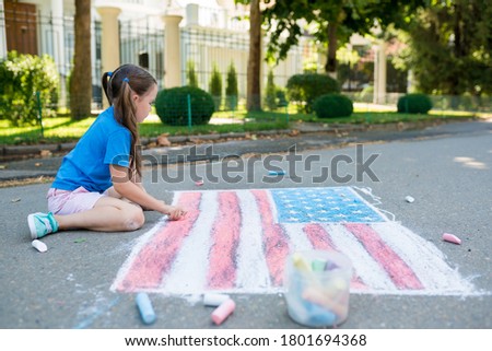 Cute  toddler girl drawing American flag with colored chalks on the sidewalk near the house on sunny summer day. Kids painting outside. Creative development of children. Patriotic day concept
