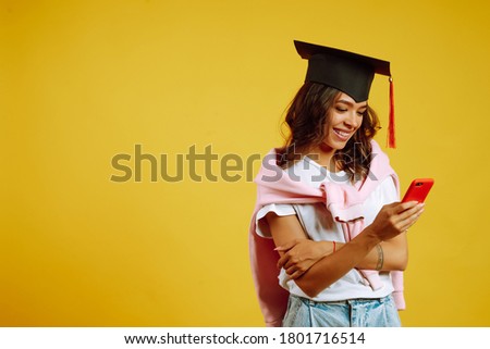 Graduate woman in a graduation hat posing with phone on a yellow background. African American woman with mobile  phone. Study, education, university, college, graduate concept.