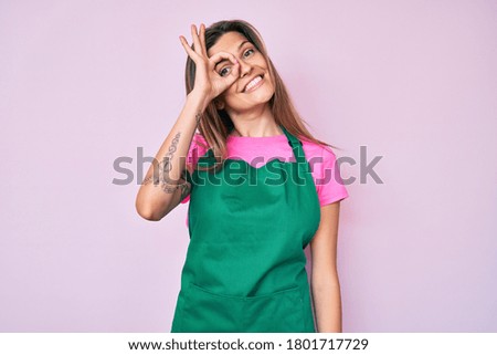 Beautiful caucasian woman wearing professional apron smiling happy doing ok sign with hand on eye looking through fingers 
