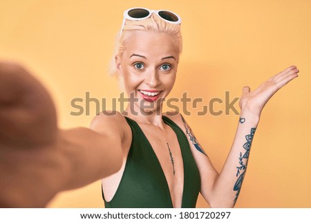 Young blonde woman with tattoo wearing swimwear taking a selfie celebrating victory with happy smile and winner expression with raised hands 