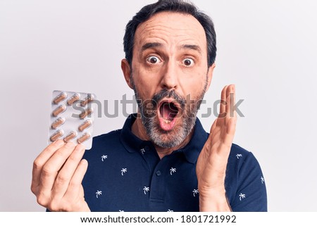 Middle age handsome man holding pills standing over isolated white background scared and amazed with open mouth for surprise, disbelief face