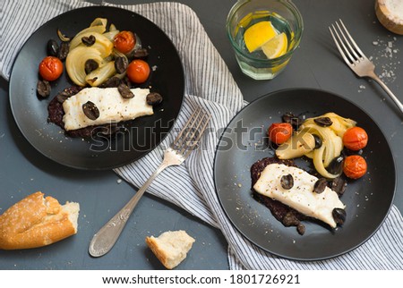 Cod with black olives and vegetables on black plate. top view