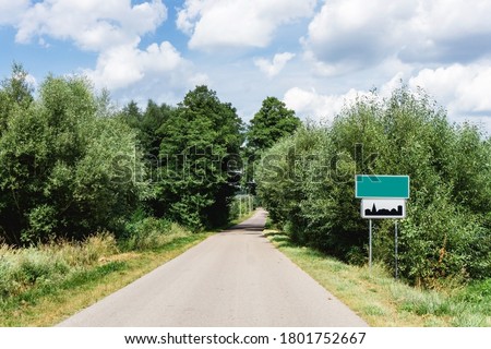 Asphalt road through the forest. Summer coniferous forest travel landscape. Long empty straight road and blue cloudy sky above. Empty copy space blank city name sign. Build up area road sign.