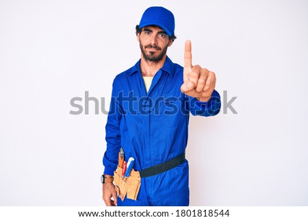 Handsome young man with curly hair and bear weaing handyman uniform pointing with finger up and angry expression, showing no gesture 