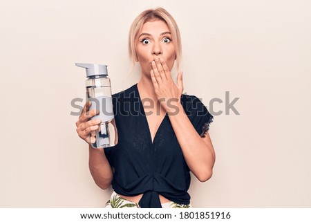 Young beautiful blonde woman drinking bottle of water over isolated white background covering mouth with hand, shocked and afraid for mistake. Surprised expression