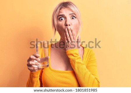 Young beautiful blonde woman drinking glass of water over isolated yellow background covering mouth with hand, shocked and afraid for mistake. Surprised expression