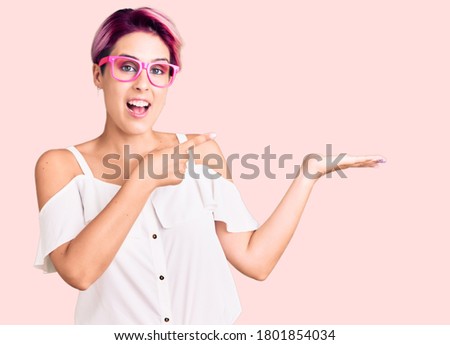 Young beautiful woman with pink hair wearing casual clothes and glasses amazed and smiling to the camera while presenting with hand and pointing with finger. 