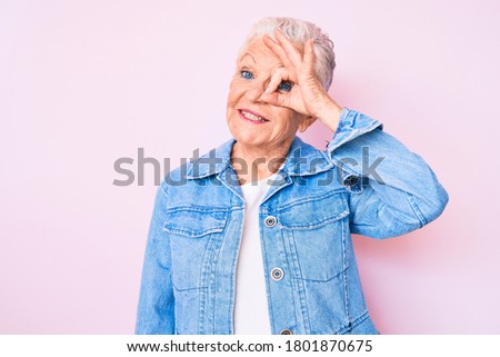 Senior beautiful woman with blue eyes and grey hair wearing casual denim jacket smiling happy doing ok sign with hand on eye looking through fingers 