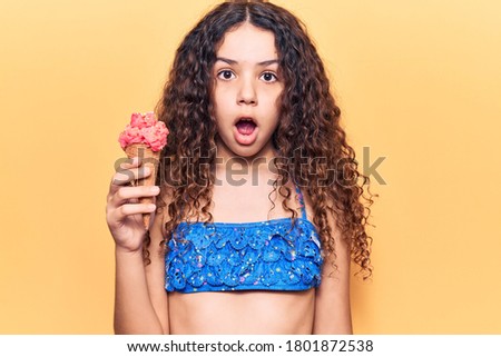 Beautiful kid girl with curly hair wearing bikini holding ice cream scared and amazed with open mouth for surprise, disbelief face 
