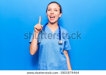 Young blonde woman wearing doctor uniform and stethoscope pointing finger up with successful idea. exited and happy. number one. 