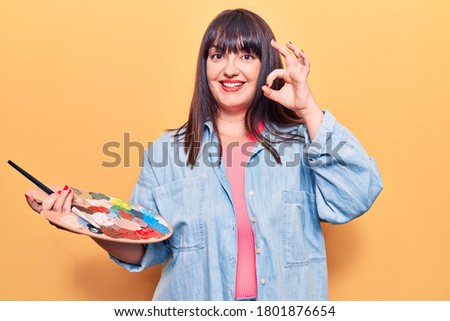 Young plus size woman holding paintbrush and palette doing ok sign with fingers, smiling friendly gesturing excellent symbol 