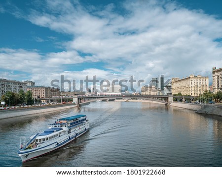 Large two-story white liner with tourists goes along Moscow river. Bridge over river in distance on background of cityscape, blue cloudy sky