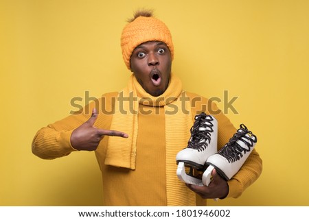 African young man in yellow hat and scarf with winter skates on yellow background.