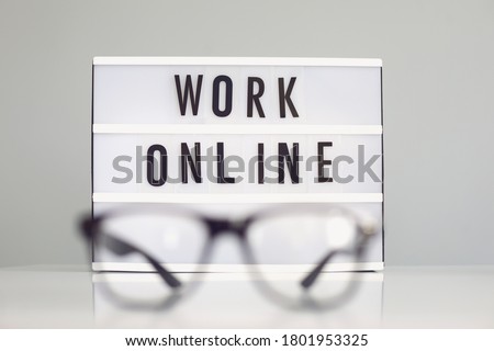 Glasses on desk and blurred sign with words WORK ONLINE against grey background. Home based work concept