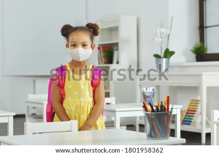an African-American girl in a yellow dress and medical mask, with a backpack on her shoulders, stands in the center of a bright classroom. back to school after lockdown and quarantine