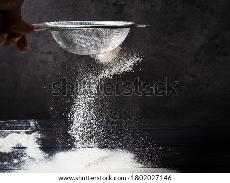 Flour is falling from the sieve. Woman hand is holding the sieve. Dark background. Close up.  Royalty-Free Stock Photo #1802027146