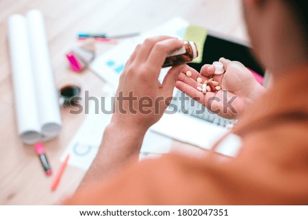 Cropped photo of a man dropping pills out of a bottle into his hand. Laptop and graphic charts on the background