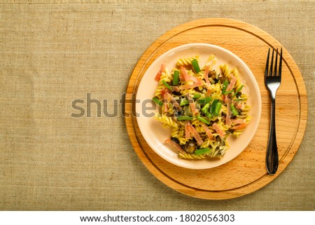 a plate with pasta with ham and mushrooms in a creamy sauce on the table with a fork on a stand on the tablecloth.