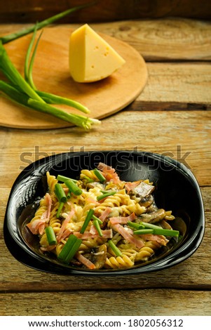 Black plate with pasta with ham and mushrooms in a creamy sauce on the table with a fork on the board.
