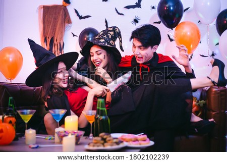 Three Happy friends sitting on sofa and wearing Halloween costumes having fun with party at nightclub or home. Group of young Asian people enjoying Halloween party with many drink and sweets. 