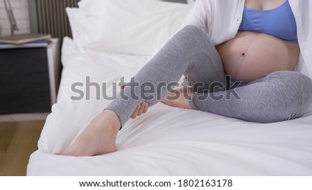 close up mother-to-be rubbing her swelling leg with hands on white bedding.