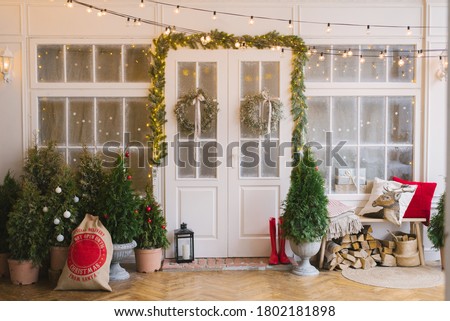 The white private house is decorated with small Christmas trees and lanterns, a bag of gifts. A bench with pillows near the front door of the house and wood under it Royalty-Free Stock Photo #1802181898