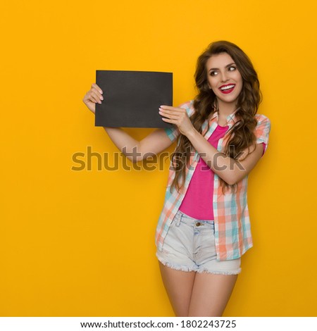 Beautiful young woman is standing with black sheet of paper and looking at it. Front view. Three quarter length studio shot on yellow background.